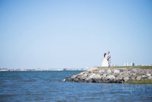 bride and groom standing on green by rocks and bay